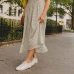 Kobieta w jasnej sukience i białych sneakersach spaceruje po miejskiej ulicy